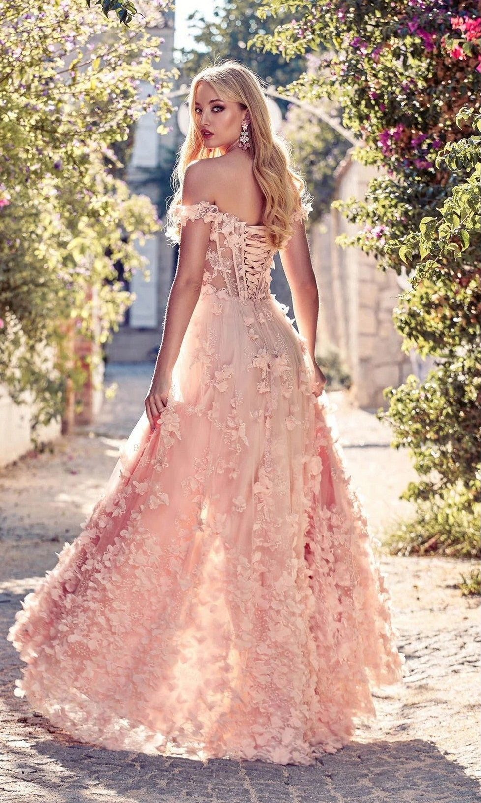 A woman wears the Off-Shoulder Long Floral Dress: Alyce 61308, featuring blush pink floral details, as she stands on a sunlit stone path surrounded by blooming plants.